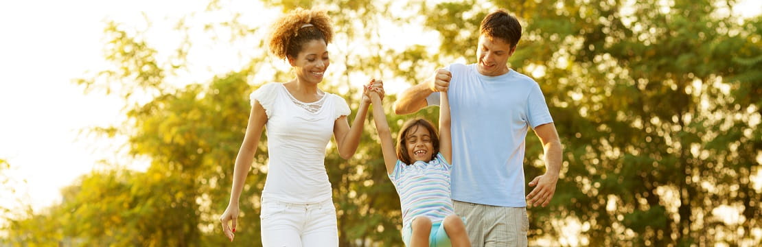 Family of three walks together. The two adults hold a young child's hands and let her swing between them while she laughs.