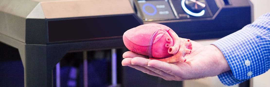A hand holds a 3D printed heart in front of a 3D printer.